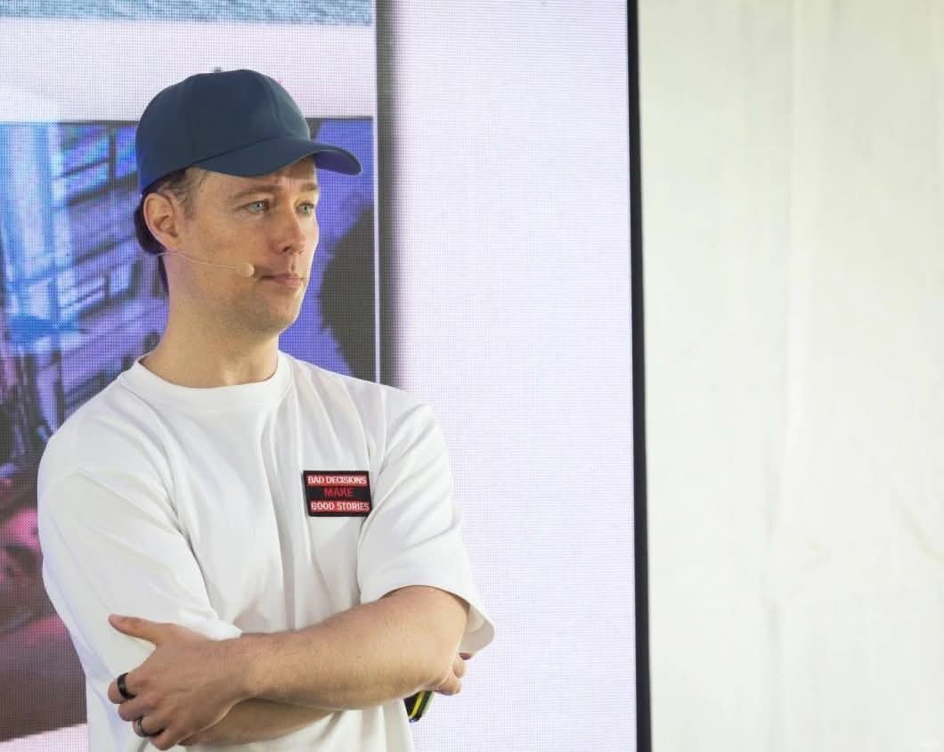 Martynas Kairys Investuotojų festivalio 2026 scenoje, sukryžiavęs rankas, su kepure ir baltais marškinėliais