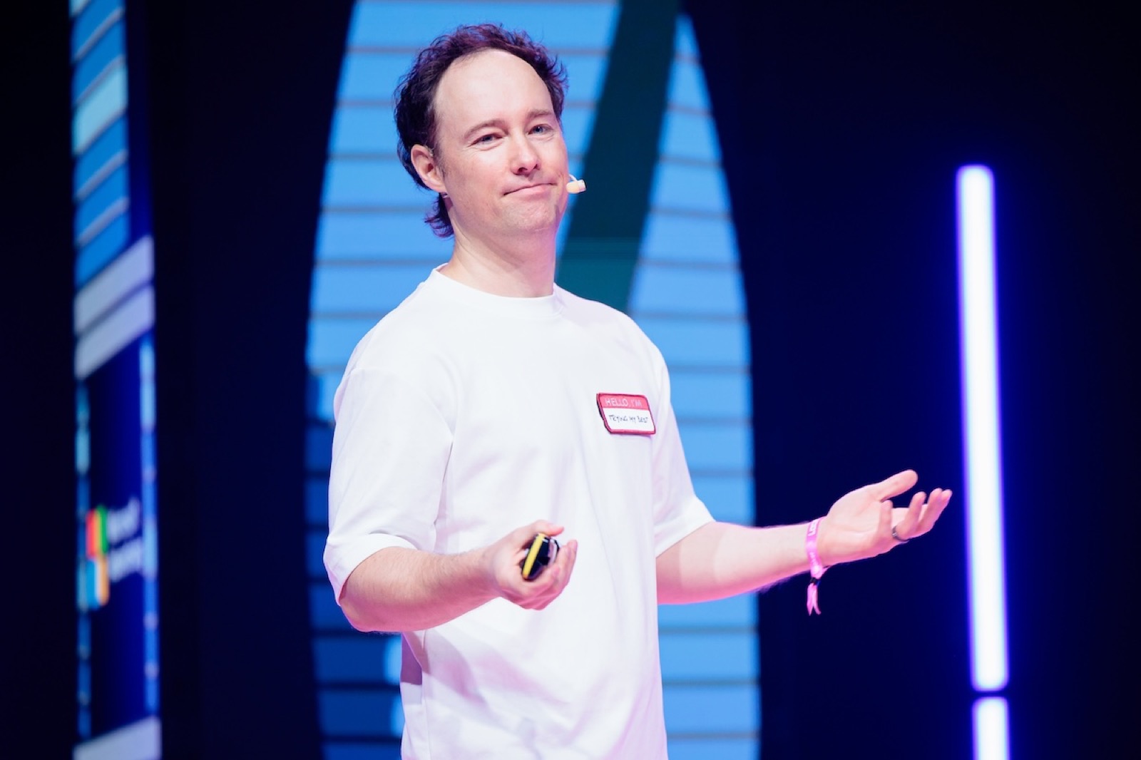 Martynas Kairys on the LOGIN 2026 stage, headset mic, mid-gesture with a name tag reading 'trying my best'
