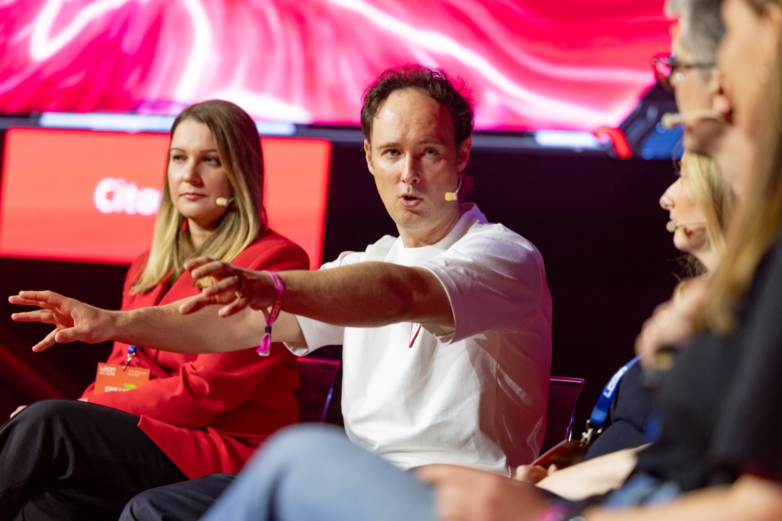 Martynas Kairys mid-answer on a LOGIN panel discussion, arm extended, seated next to another speaker in red